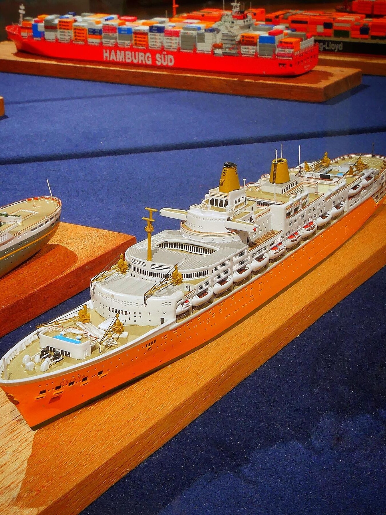 Modell eines orange-weißen Passagierschiffs mit Rettungsbooten und Deckdetails auf Holzsockel, im Hintergrund rotes Containerschiff mit Aufschrift HAMBURG SÜD.