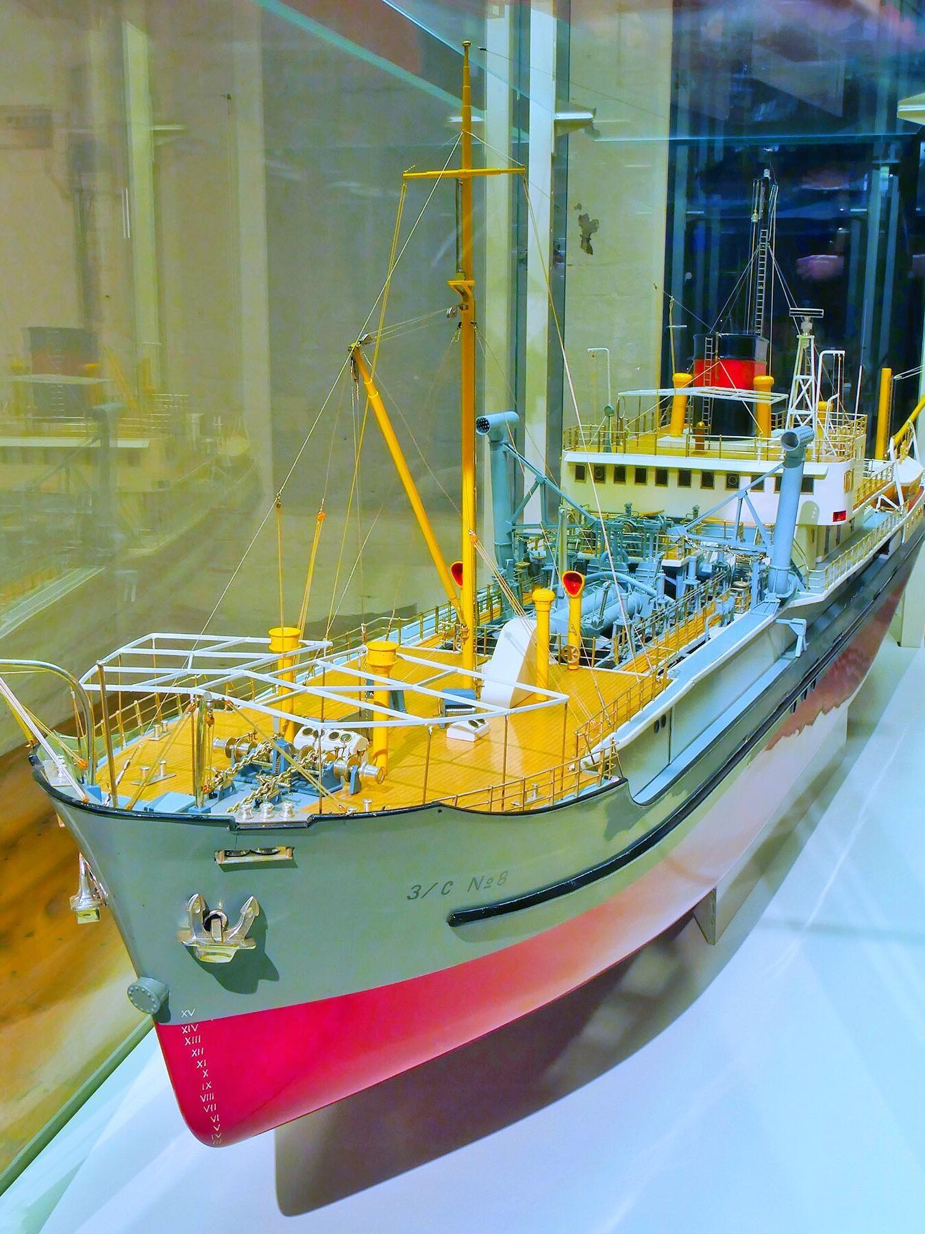 Detailliertes Schiffsmodell in Vitrine mit gelbem Deck, Masten, Takelage und rotem Unterwasserbereich, von schräg vorn gezeigt.