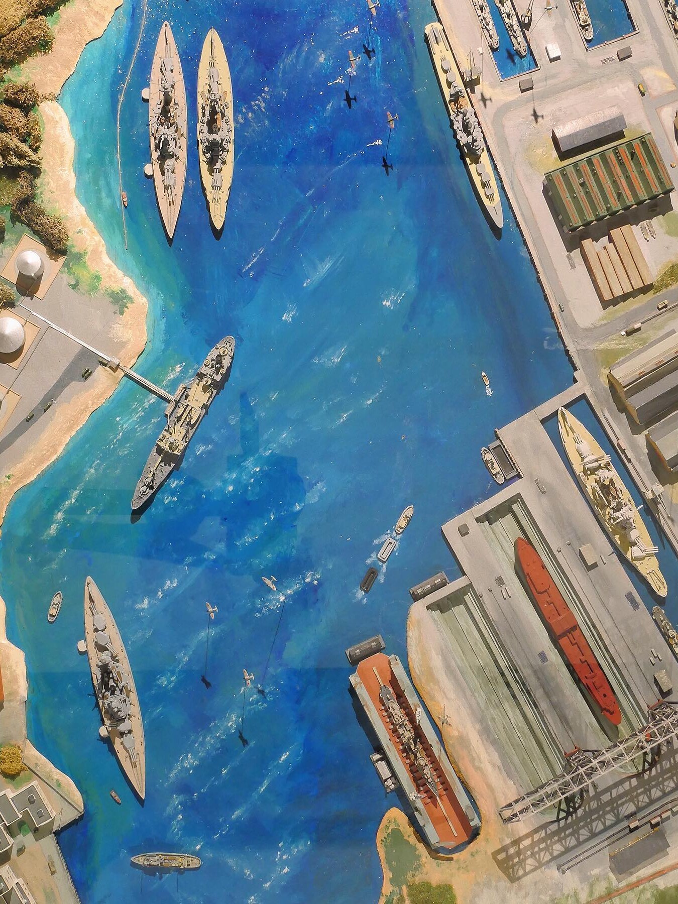 Luftaufnahme eines Hafens mit Kriegsschiffen, Frachtschiffen, Docks und kleinen Booten auf tiefblauem Wasser, umliegende Lagerhallen und Tankbehälter.