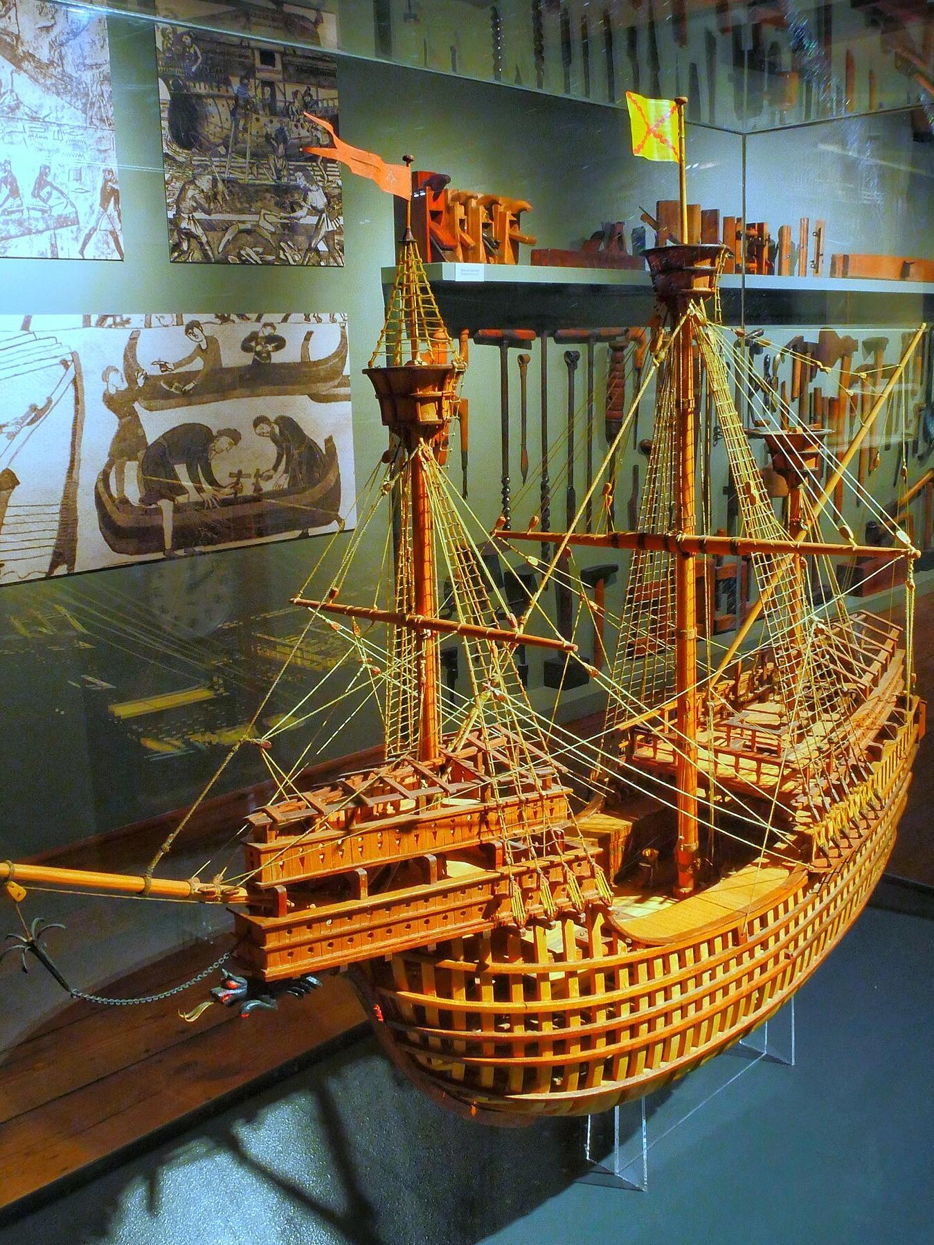 Holzmodell eines großen Segelschiffs mit Masten, Takelage und aufgesetzten Kastellen, ausgestellt vor historischen Zeichnungen.
