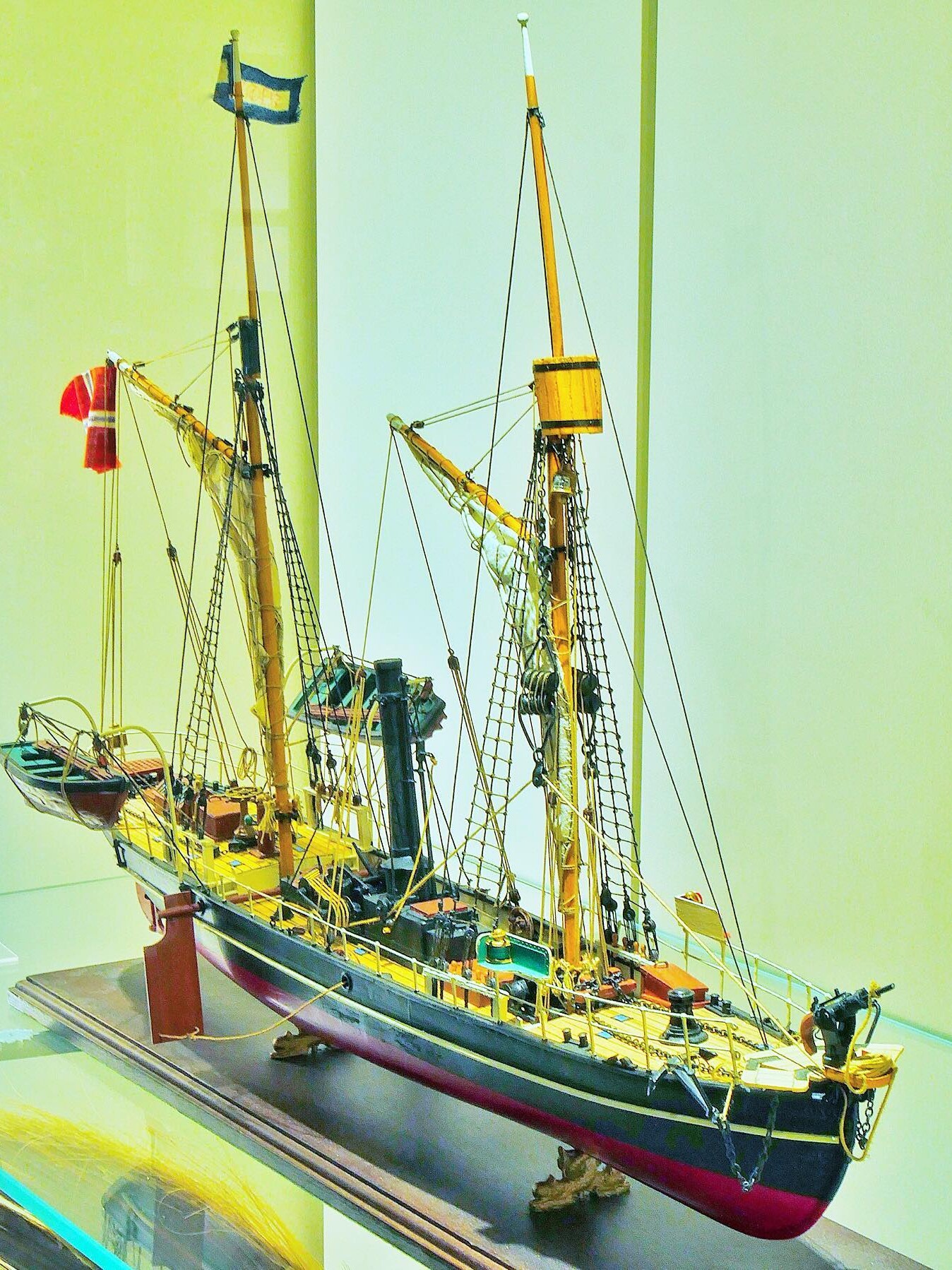 Detailliertes Modell eines historischen Segelschiffs mit zwei Masten, Takelage, Beibooten und gelb-rotem Deck in Vitrine.