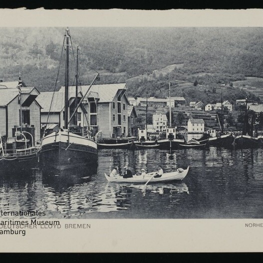 Ruderboot mit Personen vorn, mehrere angelegte Boote vor hölzernen Hafenhäusern in Norheimsund.