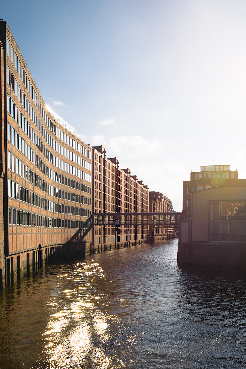 Kanal zwischen langen roten Backsteingeb&auml;uden und einem geschwungenen, verglasten B&uuml;rogeb&auml;ude, verbunden durch erh&ouml;hte &Uuml;bergangsbr&uuml;cken, mit Sonnenreflexen auf der Wasseroberfl&auml;che.