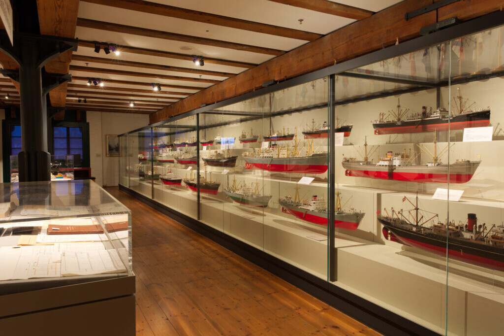 Museumsausstellung in einem Raum mit sichtbaren Deckenbalken und Holzboden; rechts lange Glasvitrinen mit mehreren detaillierten Schiffsmodellen mit roten R&uuml;mpfen, links flache Vitrinen mit Dokumenten.