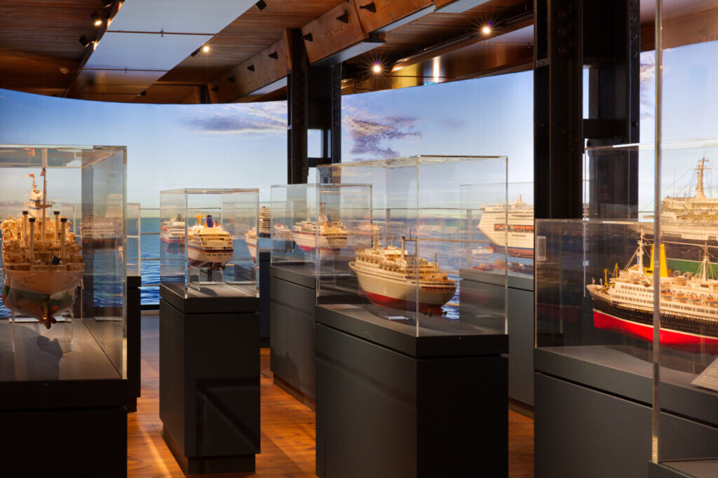 Museumsausstellung mit mehreren detaillierten Schiffsmodellen in Glasvitrinen auf dunklen Sockeln, Holzfu&szlig;boden und -decke sowie einem gro&szlig;formatigen Panoramahintergrund mit Meer und Himmel.