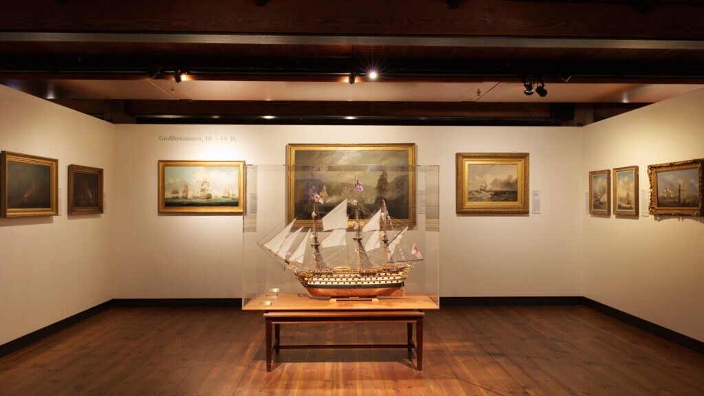Modell eines historischen Segelschiffs in einer Glasvitrine auf einem Holztisch, zentral in einem Museumssaal mit Holzfu&szlig;boden und -decke, umgeben von gerahmten Seekunstgem&auml;lden an den W&auml;nden.