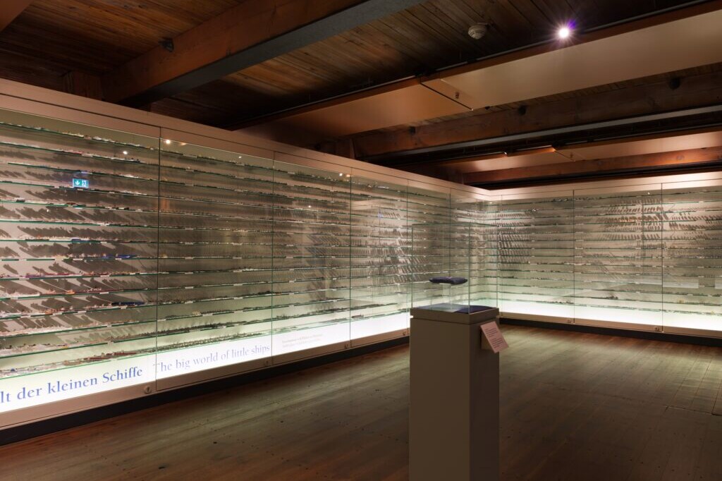 Museumsausstellungsraum mit Holzboden und -decke, beleuchteten Glasvitrinen an den Wänden, die zahlreiche kleine Schiffsmodelle auf Regalböden zeigen, und einem einzelnen Podest mit einem ausgestellten Modell in der Mitte.