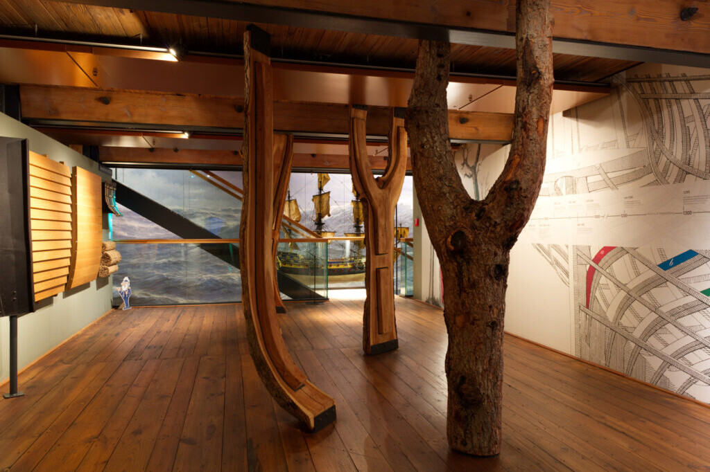 Innenansicht eines Ausstellungsraums mit sichtbaren Holzbalken, zwei gebogenen Schiffsrippen und einem vertikalen Baumstamm als Säule, im Hintergrund ein Modellsegelschiff vor einer Meereswandmalerei und technischen Zeichnungen an der Wand.